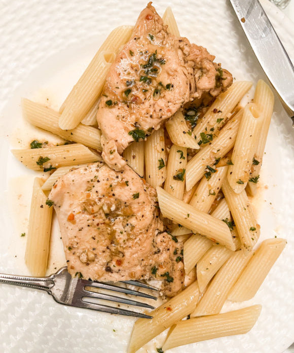 chicken with garlic butter and parsley over pasta on white plate with silver fork and knife