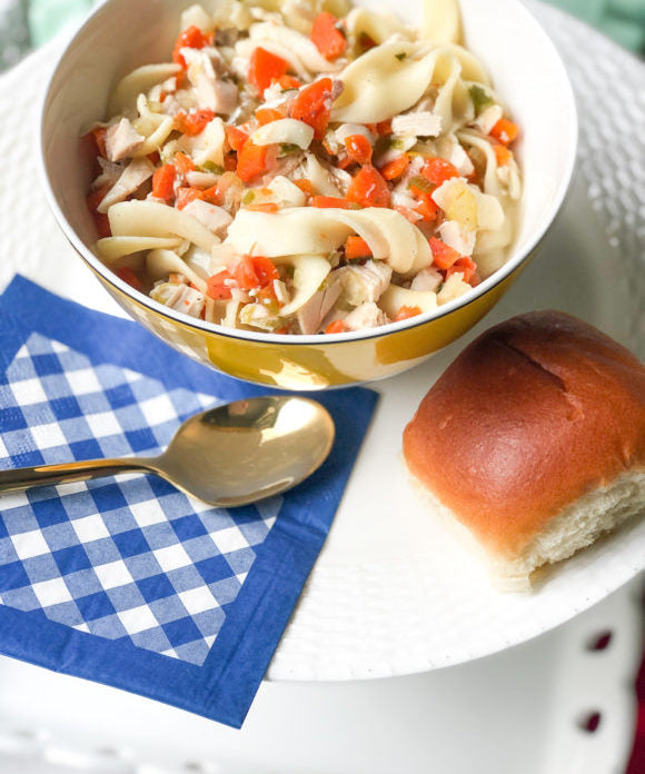chicken noodle soup with egg noodles and carrots in yellow bowl with blue and shite gingham napkin with spoon and roll