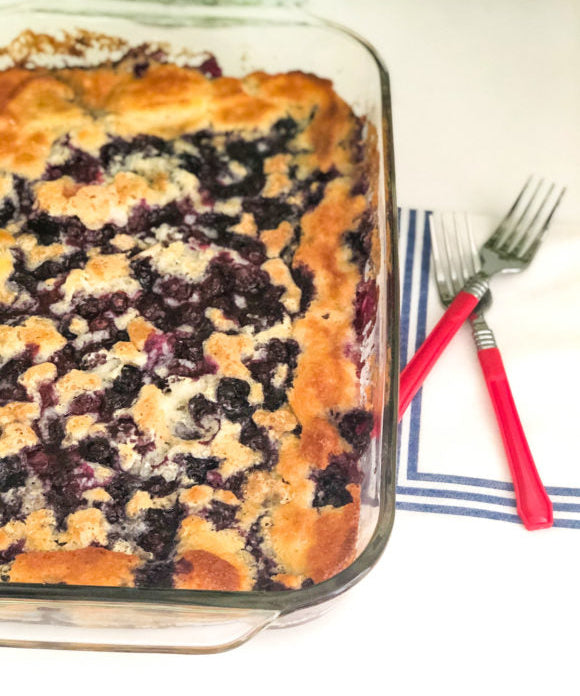 blueberry cobbler with two red forks beside glass pan on shite napkin with blue stripes