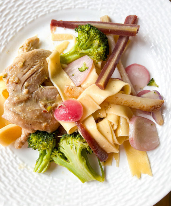 Chicken thigh on white plate served with egg noodles broccoli purple carrots and radishes