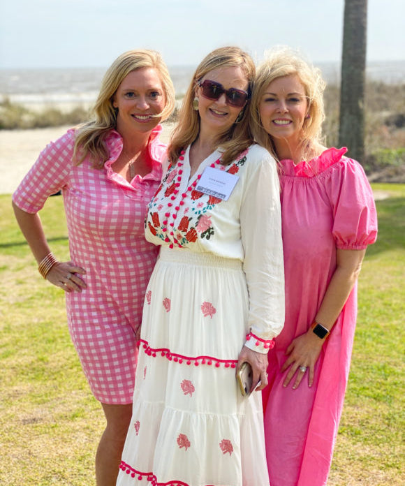 three blonde ladies standing in grass beside beach wearing pink and white dresses