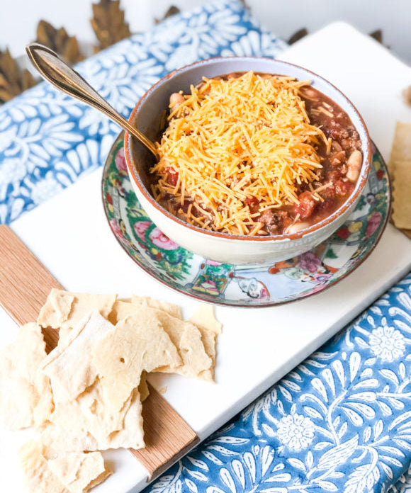 chili in a bowl with cheese on top served with crackers on a blue tablecloth and white board