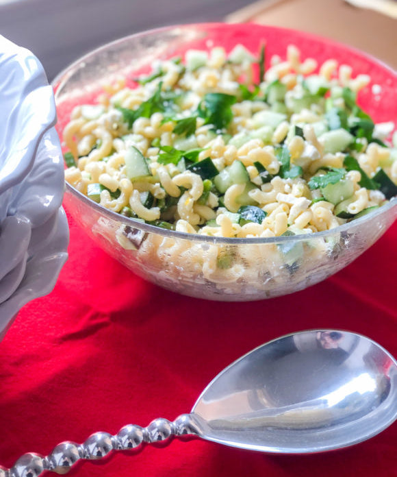 pasta salad in large clear bowl on red napkin with large silver serving spoon