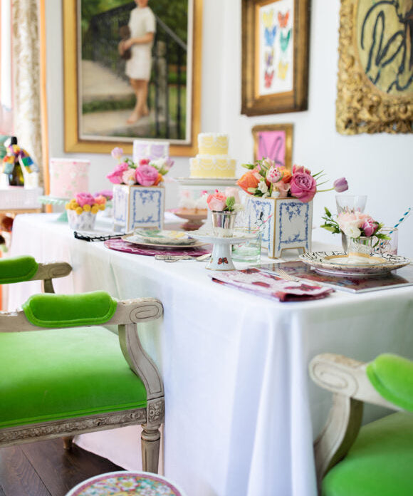table set for mother's day with green chairs flowers and cakes with art on the wall behind table