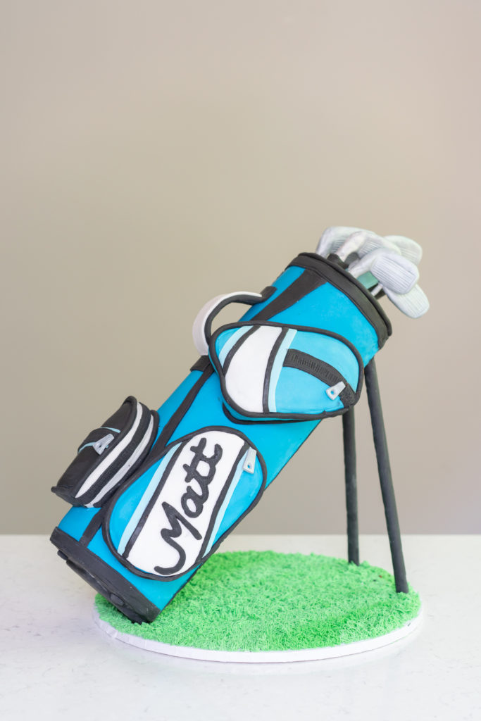 3 D golf bag cake in blue standing upright with clubs in the bag and green grass below