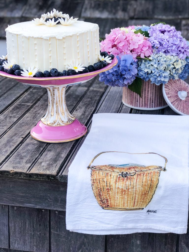 Cake with fondant daisies on pink cake stand with nantucket basket dish towel and ice bucket with hydrangeas