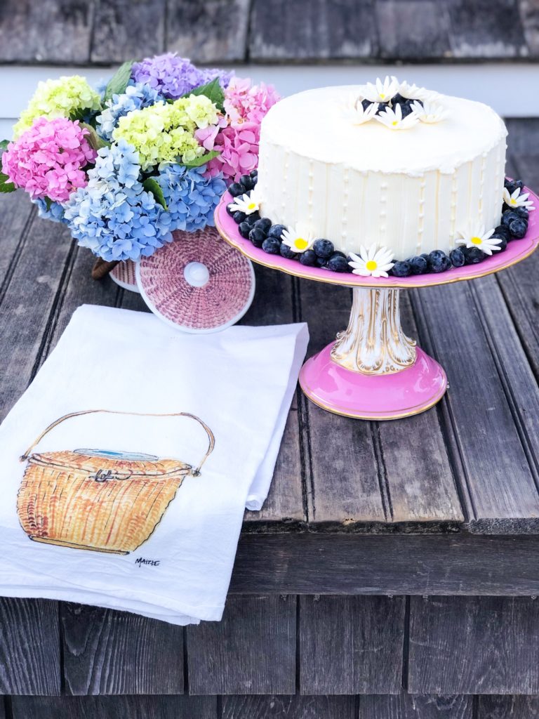 white cake with daisies on vintage pink porcelain cake stand with hydrangeas in nantucket basket and with bar towel