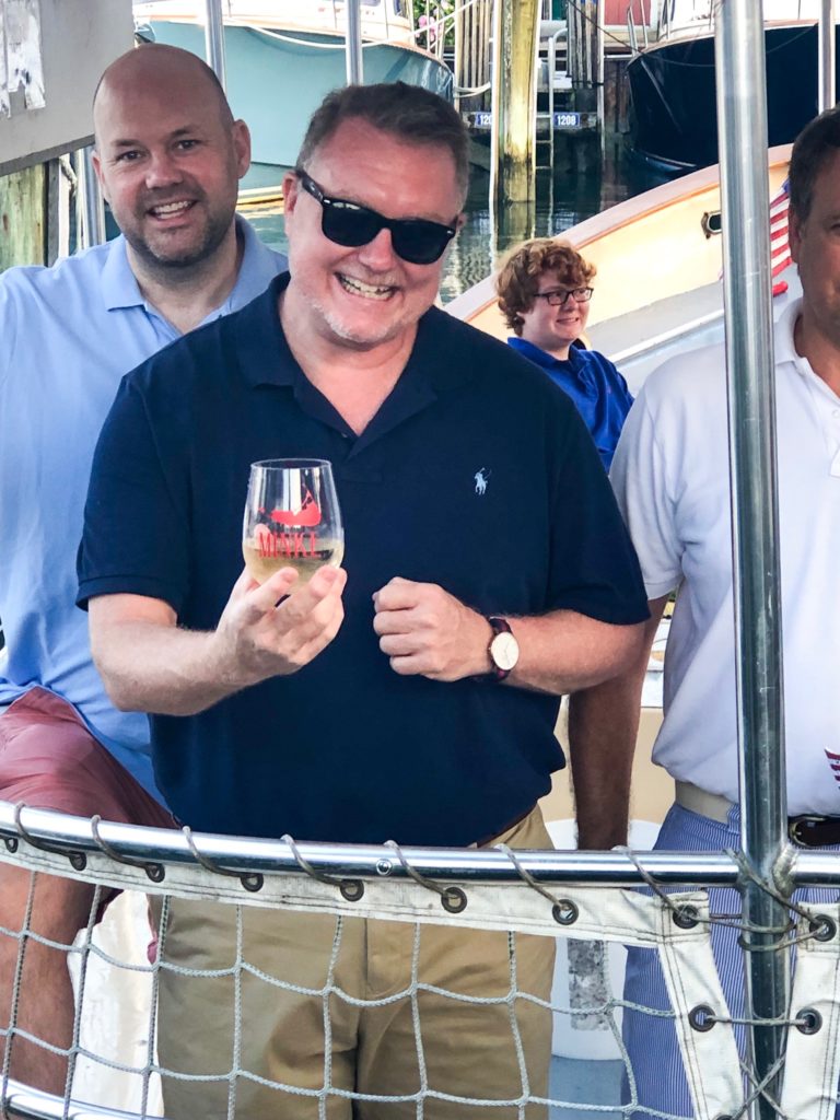 man on boat with sunglasses and navy blue shirt smiling and holding stemless wineglass