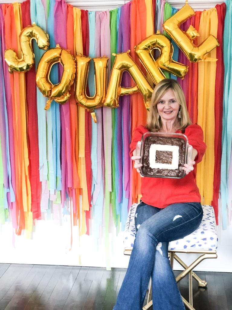 Lady in red shirt and blue jeans holding pan of brownies with square store logo and balloon letters spelling square