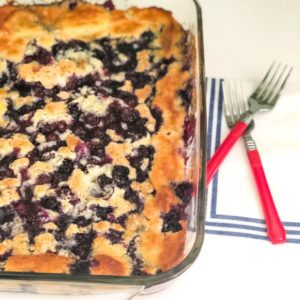blueberry cobbler with two red forks beside glass pan on shite napkin with blue stripes