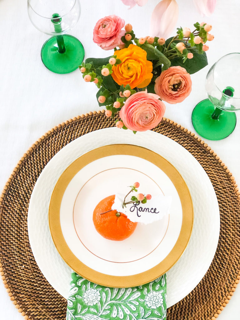 table setting overhead shot with coral colored ranuncula flowers glasses with green stems and an orange in the center of plate with name Lance written on it