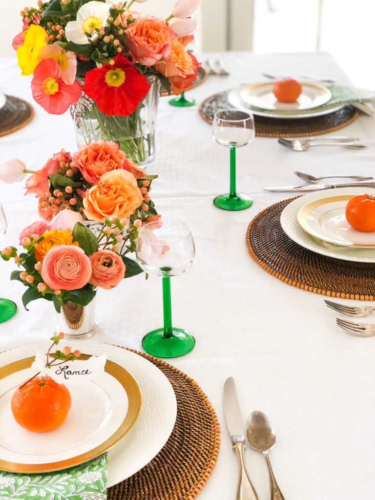 table with white table cloth brown placemats green stemmed glasses orange flowers and an orange with name on it