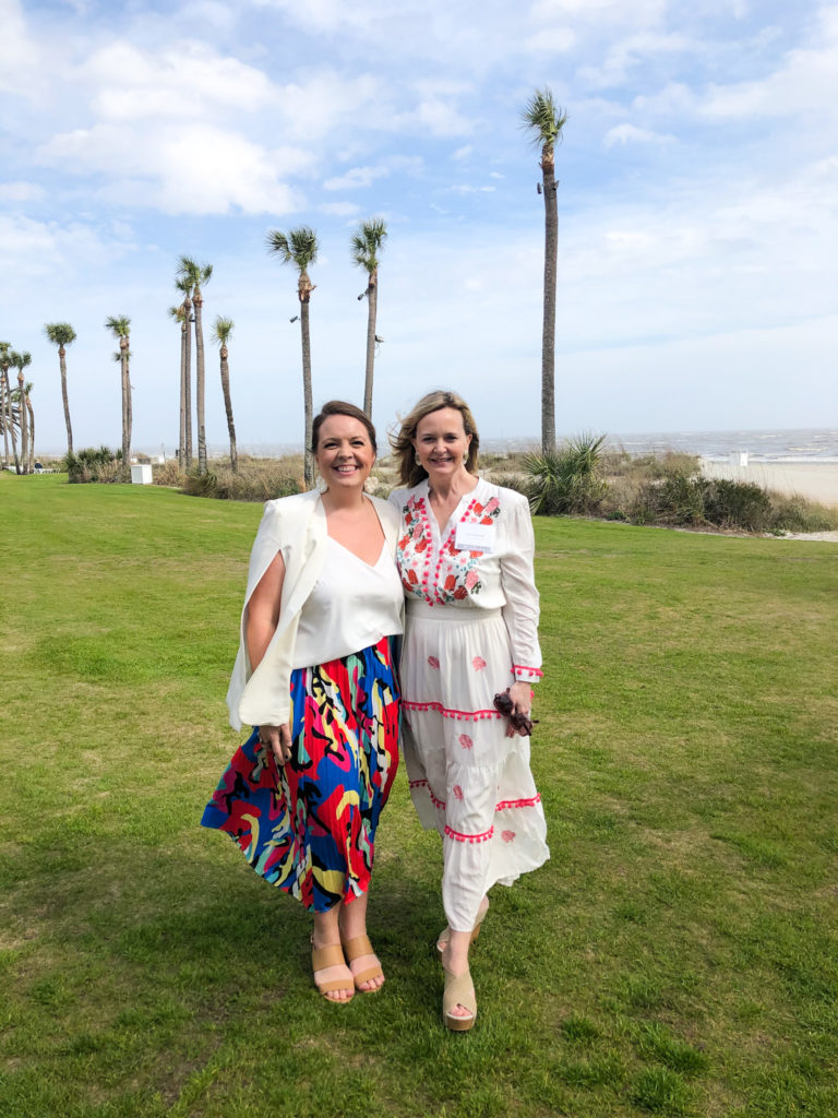 two ladies wearing dresses standing on the grass besid palm trees and the beach