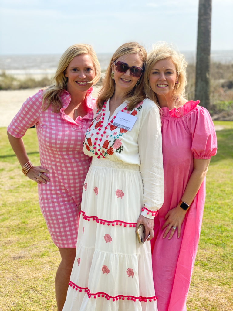 three blonde ladies standing in grass beside beach wearing pink and white dresses