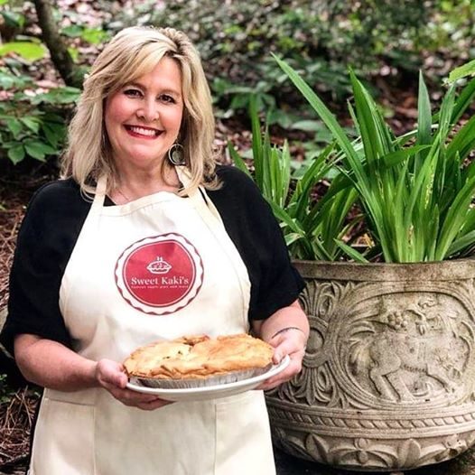 blonde lady wearing a black shirt and an ecru apron with sweet kakis on it andholding apple pie