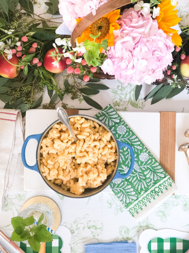 macaroni and cheese in blue pot on white cutting board and green napkin surrounded by flowers