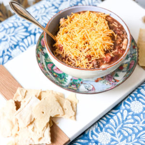 chili in a bowl with cheese on top served with crackers on a blue tablecloth and white board