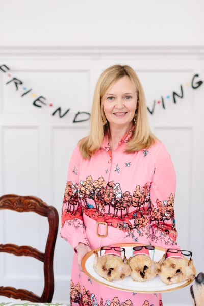 lydia menzies in pink printed dress holding cornish hens on a platter in front of friendsgiving banner