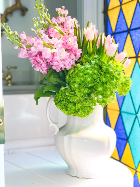 pink and green flowers in a white pitcher beside stain glass window