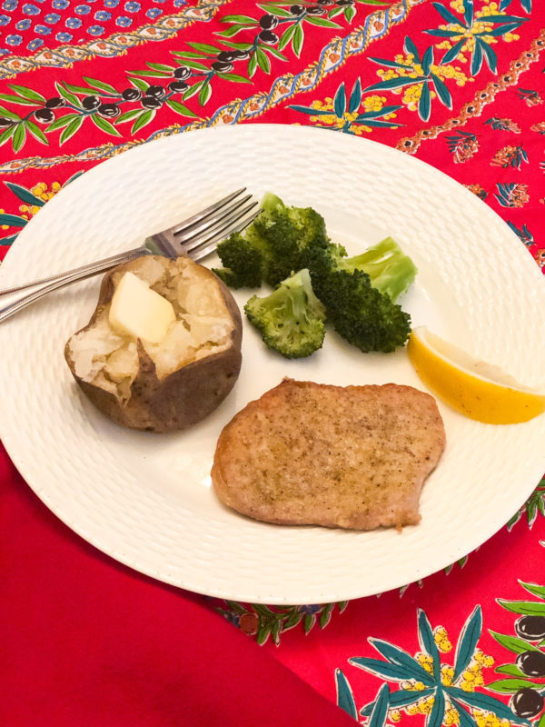 pork chop baked potato broccoli and lemon on white plate and red floral tablecloth