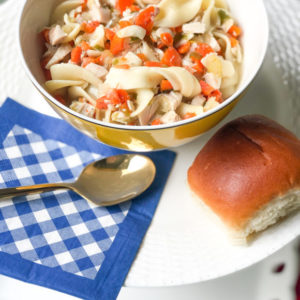 chicken noodle soup with egg noodles and carrots in yellow bowl with blue and shite gingham napkin with spoon and roll