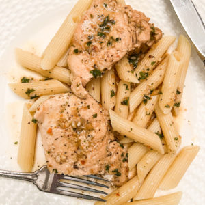 chicken with garlic butter and parsley over pasta on white plate with silver fork and knife