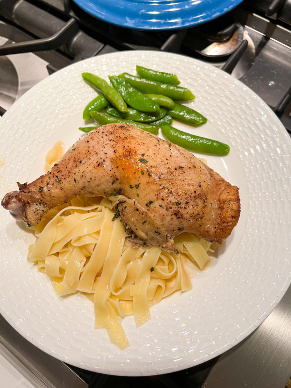 whole baked chicken leg with egg noodles and sugar snap peas on a white plate