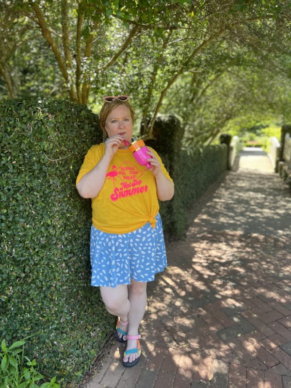 Woman wearing "School Bus Stop Ahead, Next Stop Summer" Tee with a blue skirt and sandals while drinking from a bring pink cup.