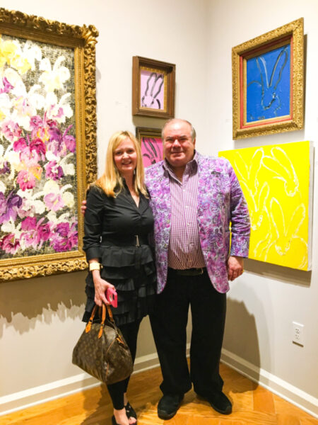 blonde lady wearing black dress standing next to artist hunt slonem with his paintings