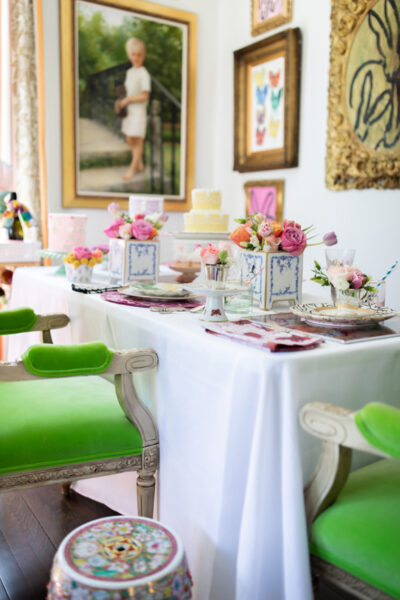 table set for mother's day with green chairs flowers and cakes with art on the wall behind table
